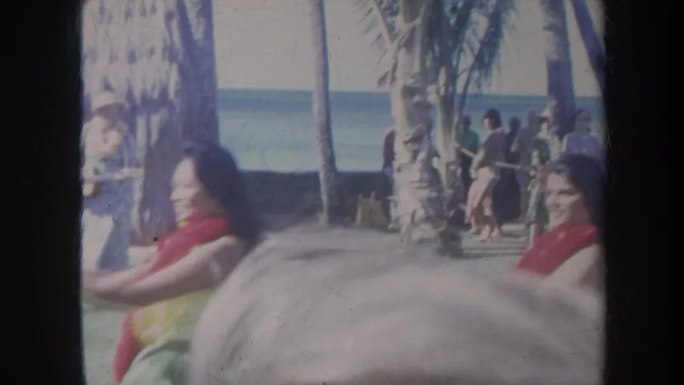 1967 Honolulu home-movie frame with dancers, spectators, musicians, palm trees, and a soft obstruction at the bottom of the frame.