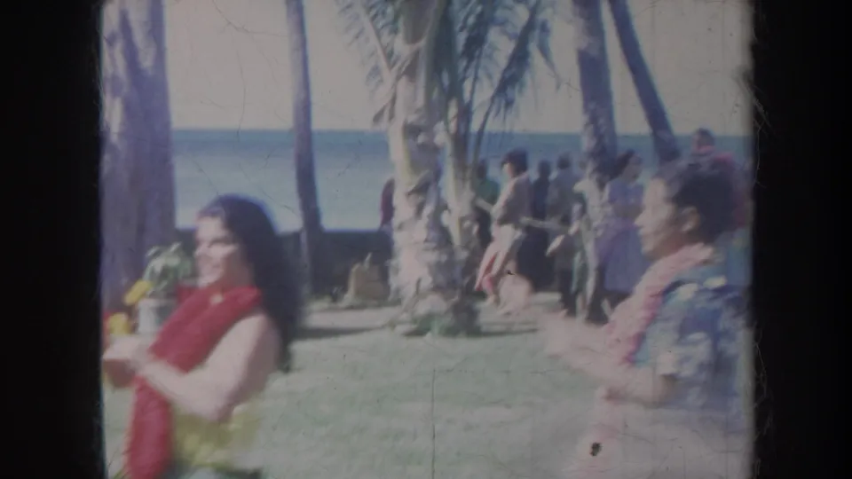 1967 Honolulu frame showing a smiling hula dancer in lei and skirt, with spectators, palm trees, and ocean in the background.