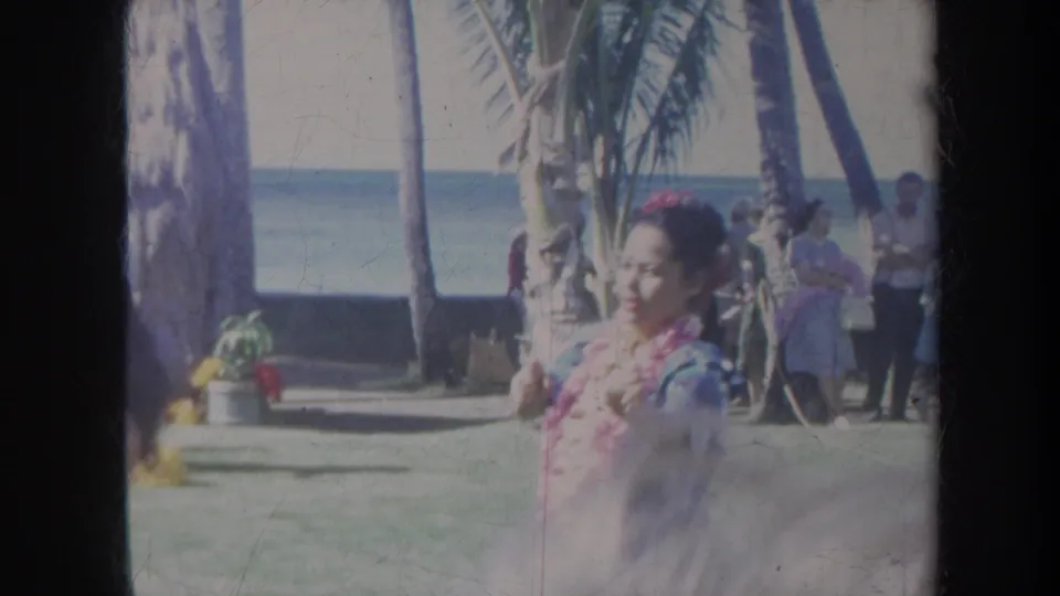 1967 Honolulu home-movie frame showing a hula dancer in red lei and green skirt near palm trees, spectators, and the ocean.