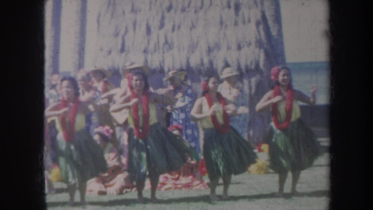 Group of hula dancers in green skirts and red leis performing before seated spectators.