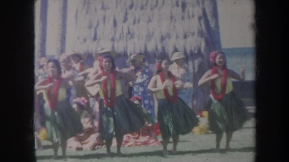 1967 Honolulu frame with several hula dancers in green skirts and red leis performing in front of an outdoor audience.