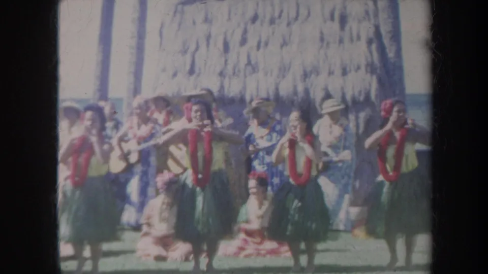 1967 Honolulu home-movie frame showing dancers in leis and skirts performing on grass before a thatched backdrop and audience.