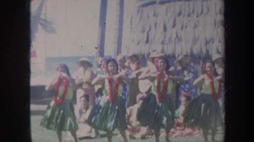 1967 Honolulu frame showing dancers in formation beside spectators, palm trees, a thatched structure, and oceanfront light.