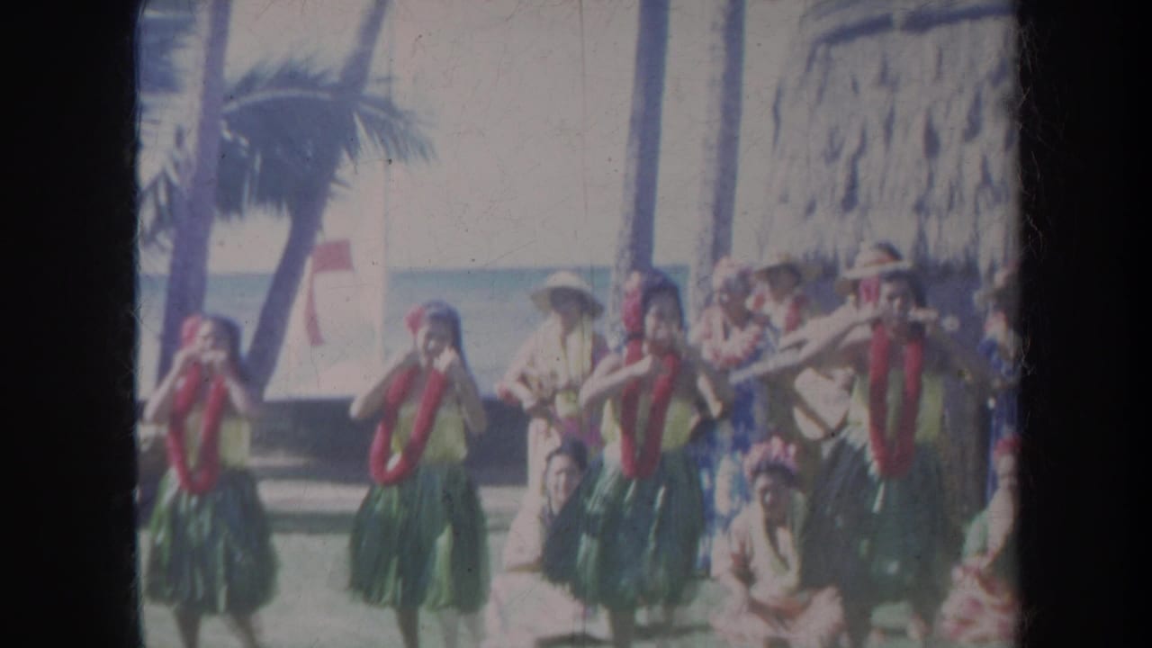 Hula dancers in green skirts and red leis perform near palm trees and the ocean.