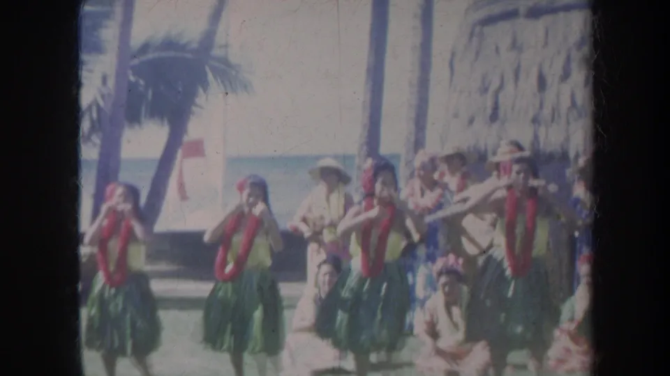 1967 Honolulu home-movie frame showing hula dancers in green skirts and red leis performing on a beach with palm trees behind them.