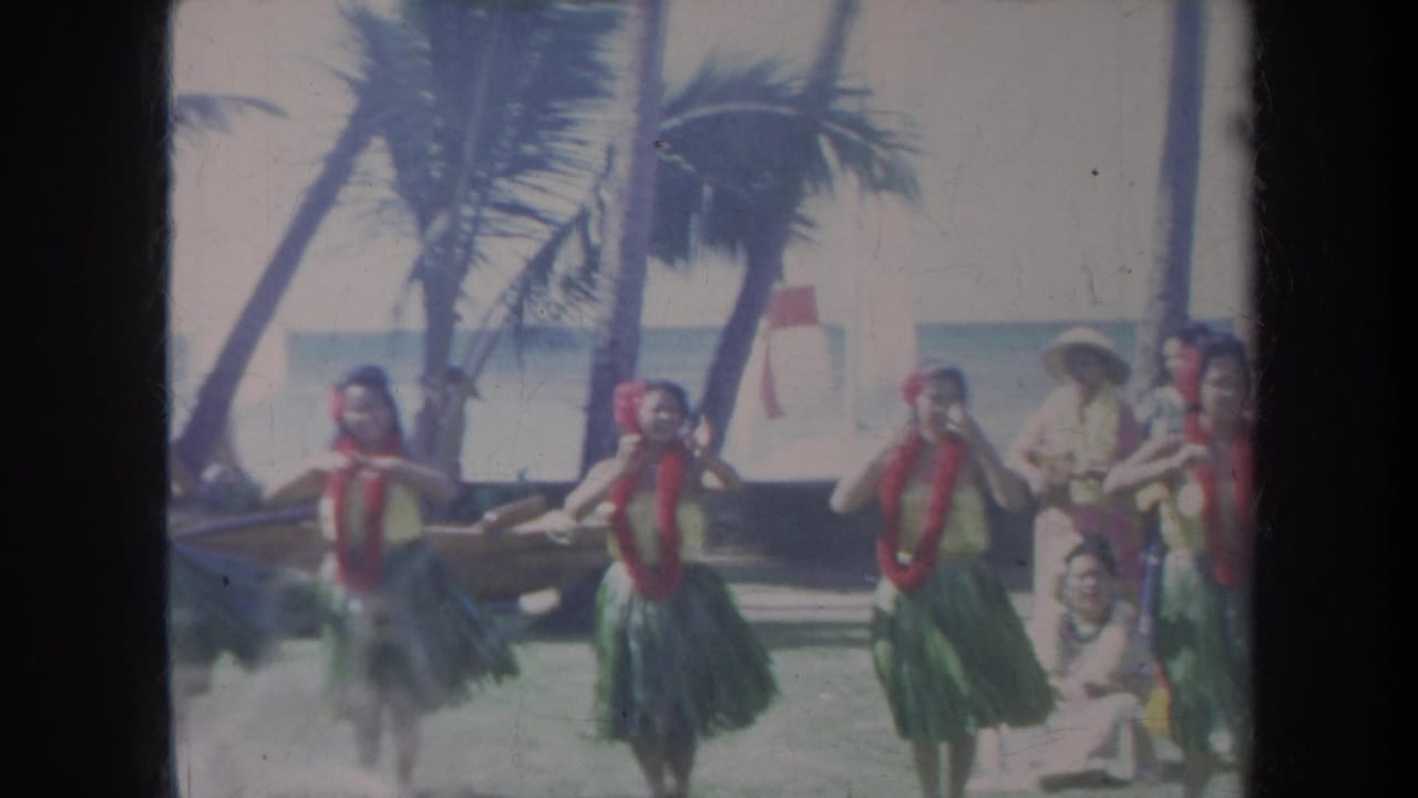 1967 Honolulu footage frame with hula dancers in leis and grass skirts moving in formation beside a beach and palm trees.