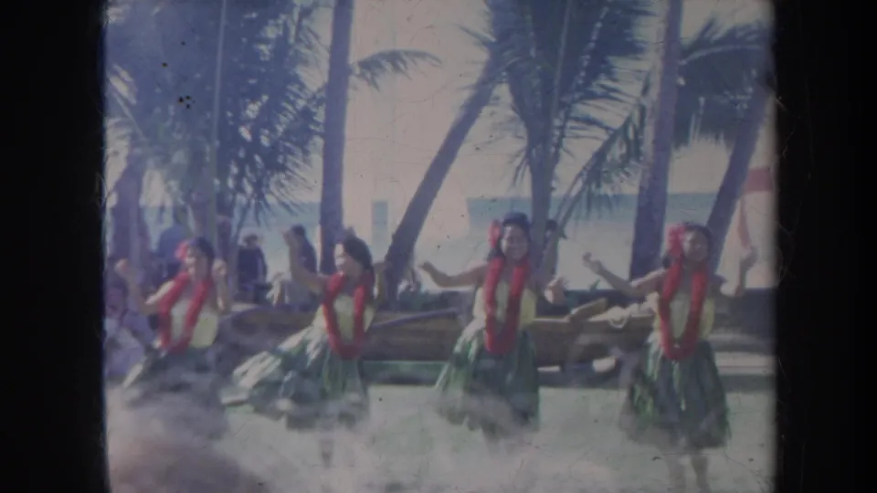 1967 Honolulu home-movie frame of hula dancers gesturing with arms extended during a palm-shaded beach performance.
