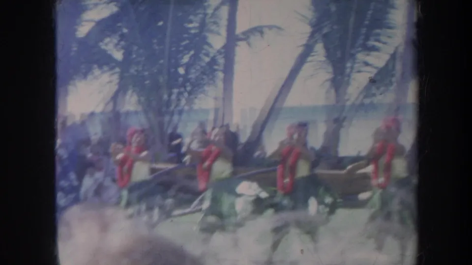 1967 Honolulu beach hula performance frame with dancers partly softened by foreground blur and palm trees behind them.