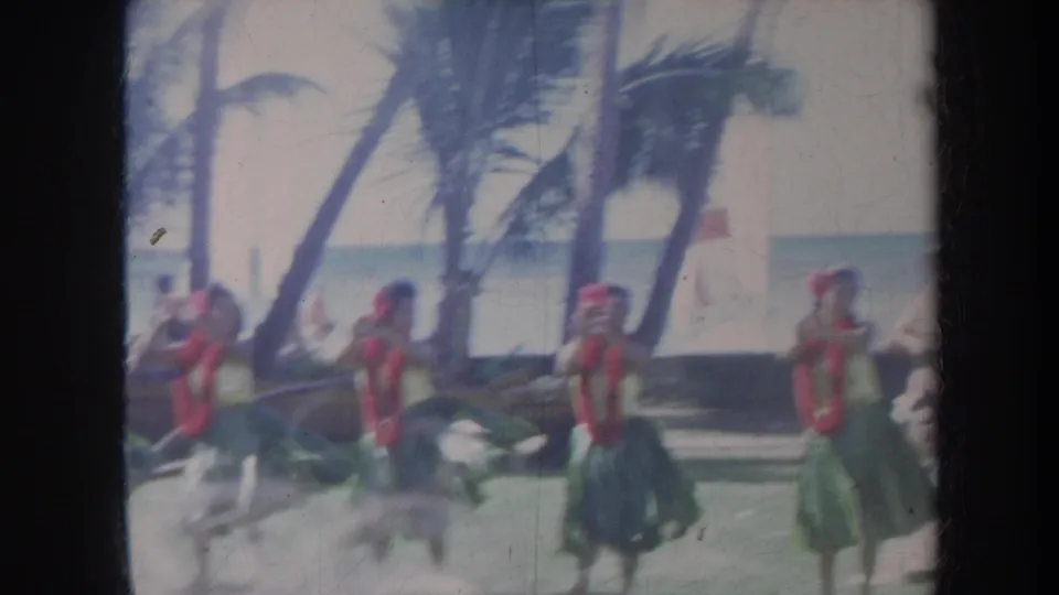 1967 Honolulu home-movie frame showing hula dancers in red leis and green skirts performing near palm trees with the ocean behind them.