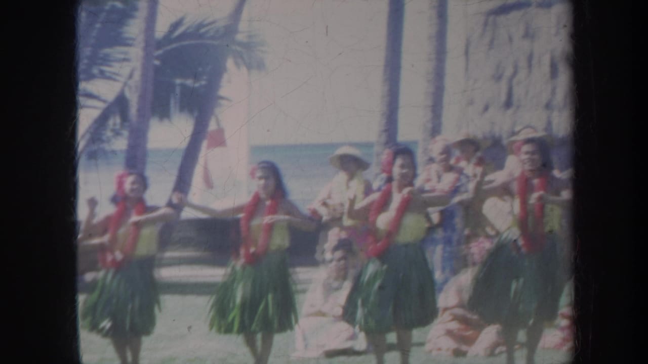 1967 Honolulu beach performance with hula dancers in leis and grass skirts, spectators behind them, and a thatched shelter at right.