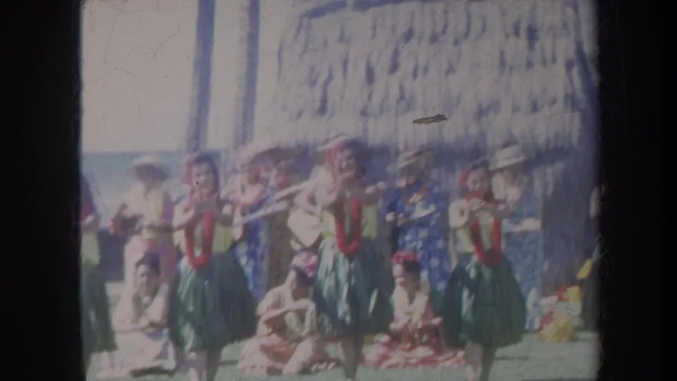 Soft 8mm frame of a Honolulu beach hula performance with dancers in leis and skirts, seated spectators, and musicians or attendees behind them.