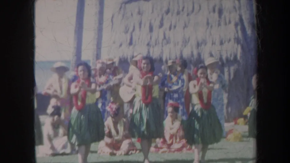 1967 Honolulu frame showing three hula dancers in red leis and green skirts performing before spectators under a thatched beach structure.