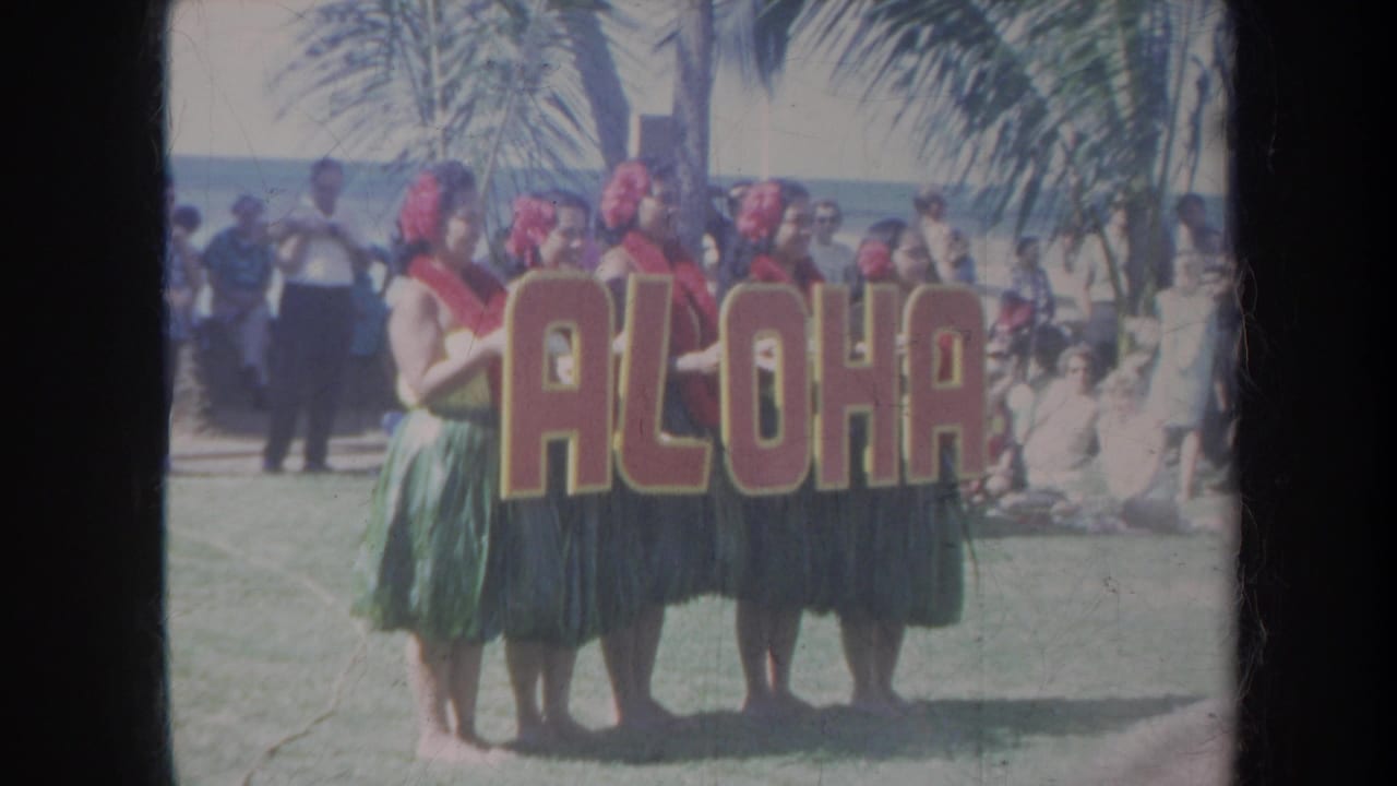 1967 Honolulu home-movie frame of five women in leis and grass skirts holding an ALOHA sign with palm trees and spectators behind them.