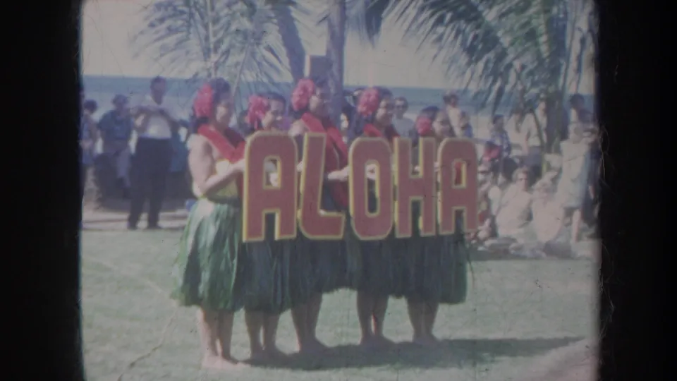 1967 Honolulu home-movie frame of five women in leis and grass skirts holding an ALOHA sign with palm trees and spectators behind them.