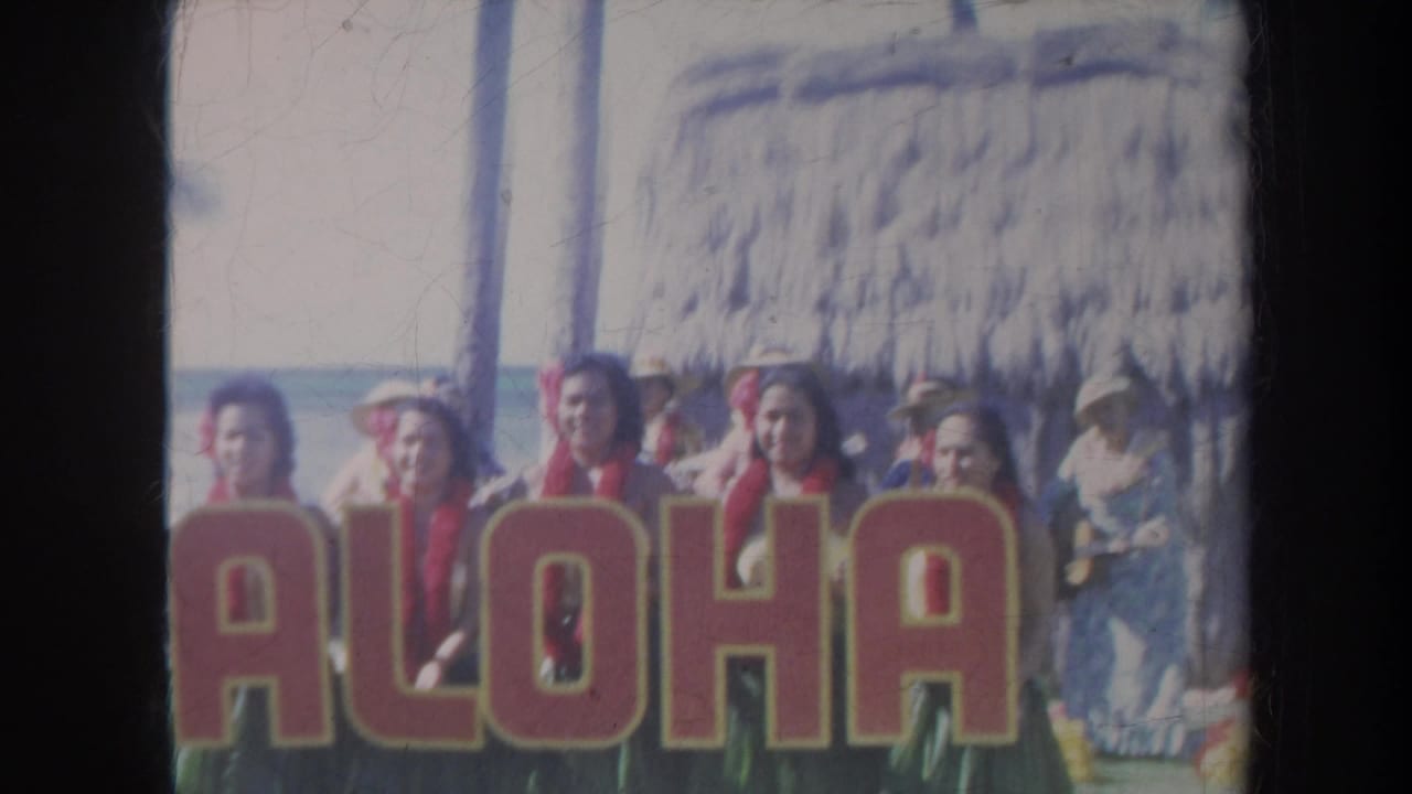 Women stand behind a large ALOHA sign beside a thatched beach shelter.