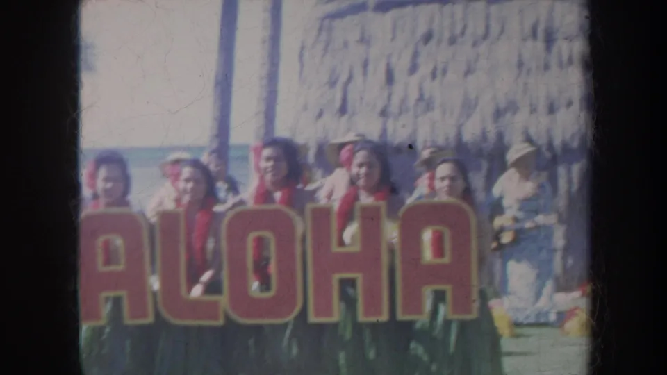 Honolulu 1967 home-movie frame with the ALOHA letters prominent, performers partly visible behind the sign, and a thatched shelter nearby.
