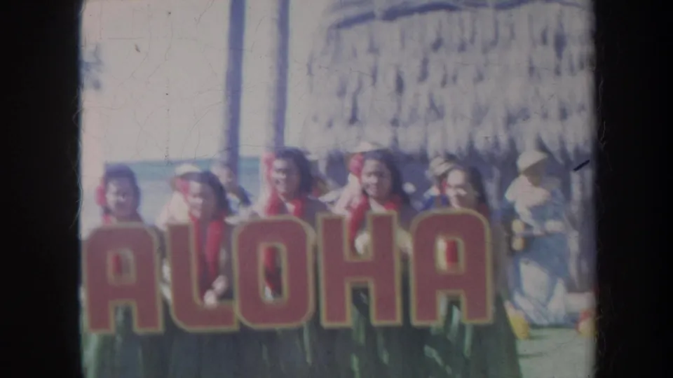1967 Honolulu home-movie frame of women behind an ALOHA sign, with beach onlookers, palm trunk, and thatched structure in the background.