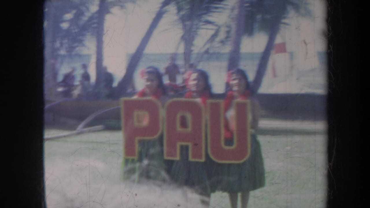 Women in grass skirts stand by a large red-letter banner near palm trees and the ocean.