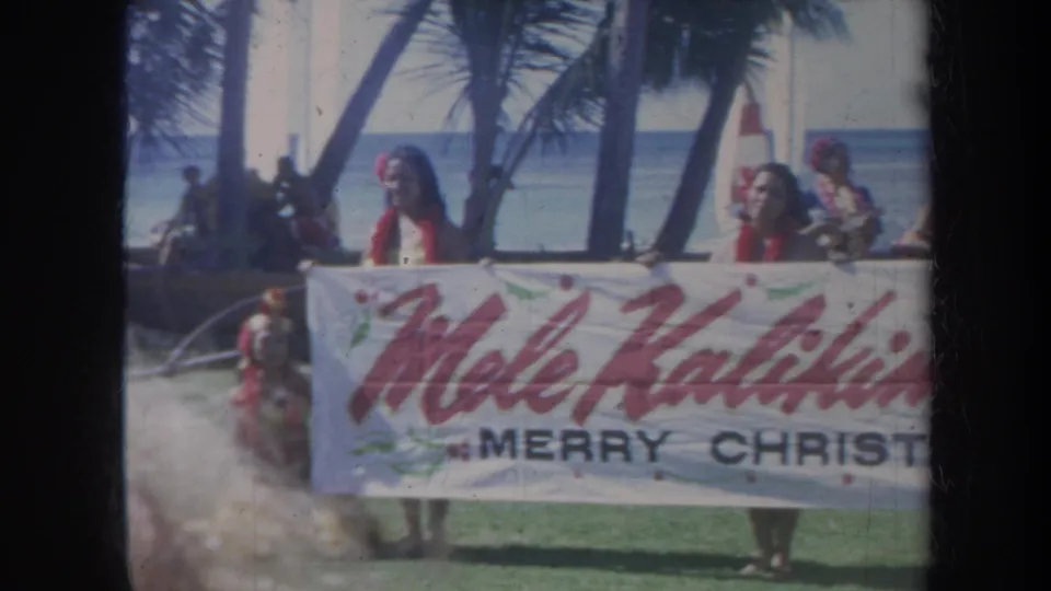 1967 Honolulu frame with women holding a Mele Kalikimaka Merry Christmas banner against a palm-lined beach and blue ocean backdrop.