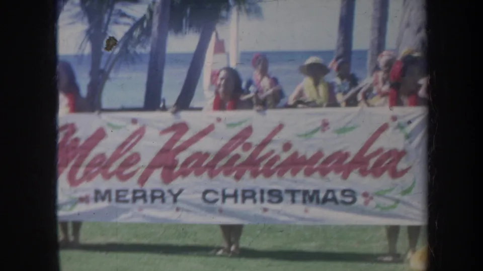 1967 Honolulu home-movie frame showing the Mele Kalikimaka banner stretched wide with people behind it on a sunny beach setting.