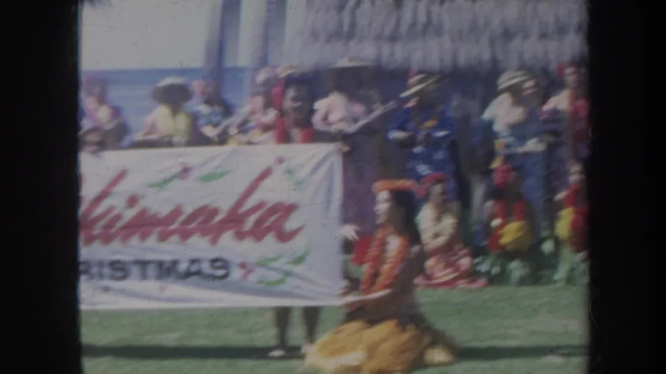 1967 Honolulu frame showing a kneeling dancer in grass skirt near the Christmas banner, with spectators, palms, ocean, and a thatched roof nearby.