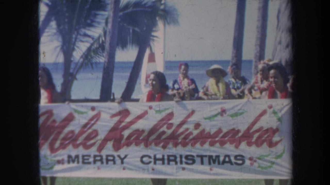 Beach crowd behind a Mele Kalikimaka Merry Christmas banner with palm trees and ocean beyond.