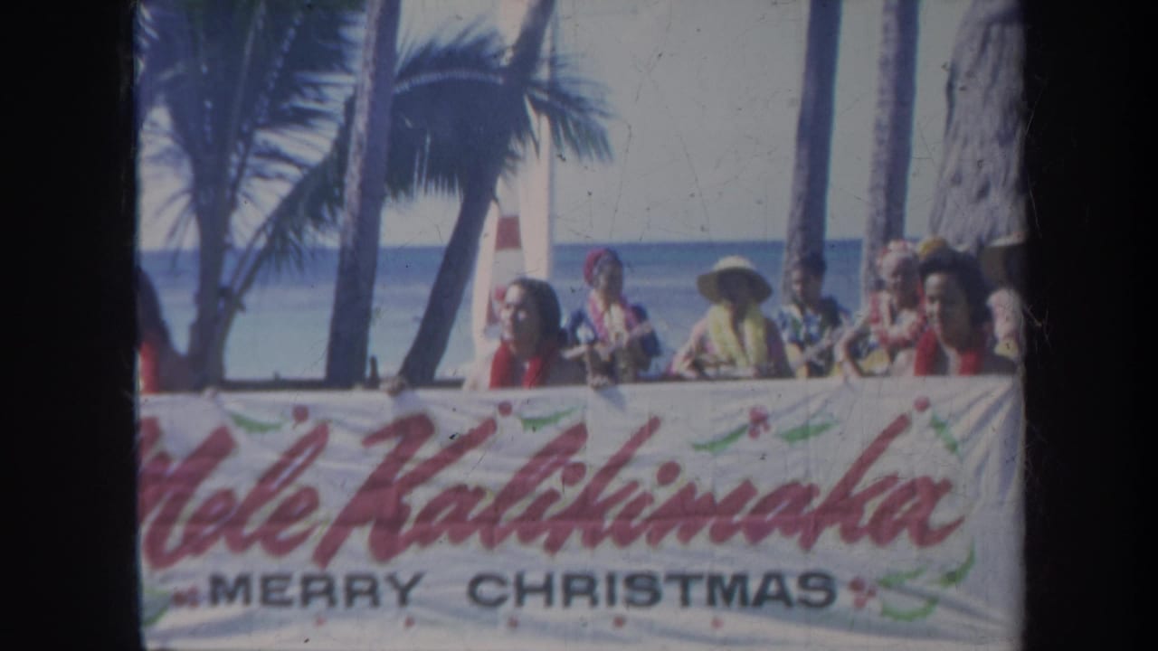 People in leis and beachwear stand behind a Christmas banner near palm trees and ocean water.