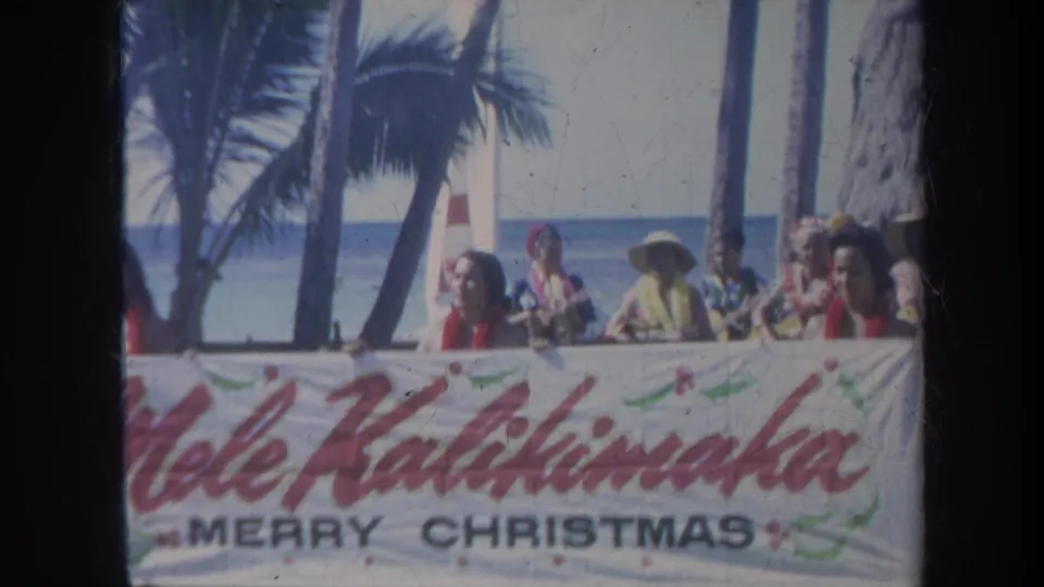 1967 Honolulu beach scene with a group behind a large Mele Kalikimaka Merry Christmas banner by the ocean.