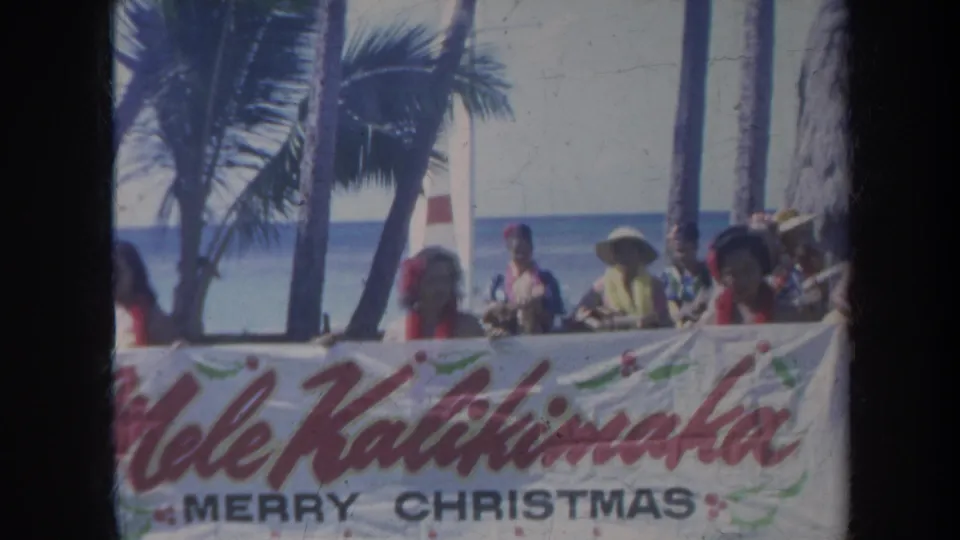 Honolulu 1967 home-movie frame with people and leis behind a red-lettered Mele Kalikimaka banner at the beach.