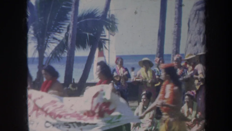 1967 Honolulu beach frame showing a holiday banner being carried or adjusted with seated and standing people nearby.