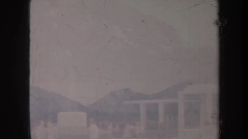 1967 Honolulu home-movie frame with a distant white structure, faint people, mountains, and heavy 8mm scratches.