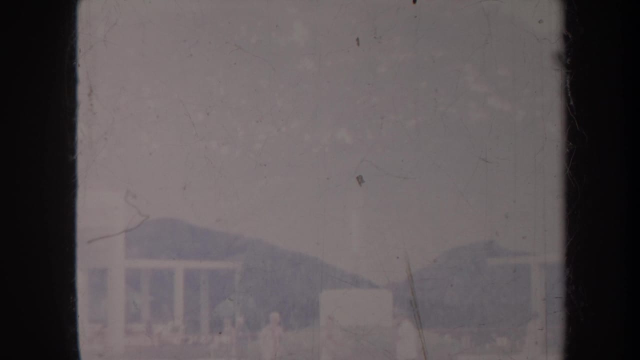 White building and mountain backdrop partly obscured by film scratches and dark frame edge.