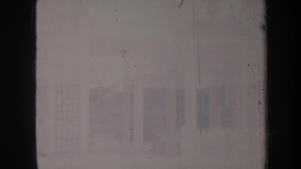 Underexposed 1967 Honolulu home-movie frame with faint single-story building shapes, hedges, and dark vertical film edges.