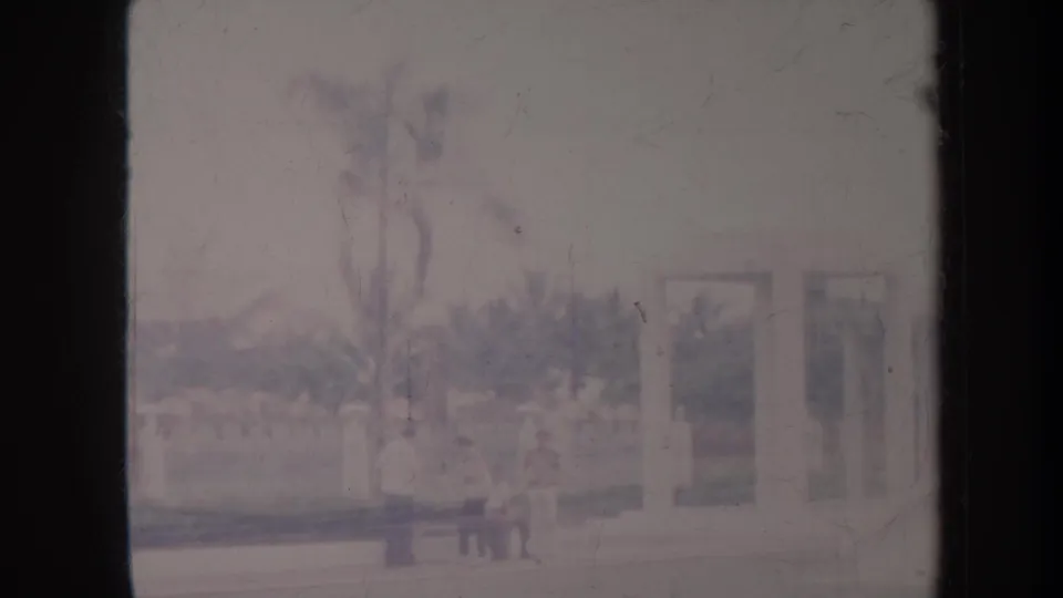Soft 1967 Honolulu frame showing landscaped grounds, a low wall, and indistinct statue-like forms near a pale building.