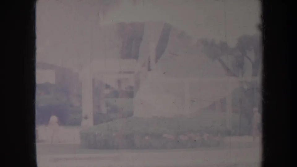 1967 Honolulu frame showing a garden walkway, trimmed hedges, a small open-sided structure, and tiny distant pedestrians in faded color.