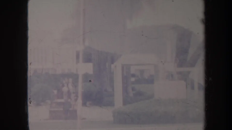 Honolulu 1967 home-movie frame with a small gazebo-like pavilion, manicured hedge, walkway, and indistinct people moving in the garden area.