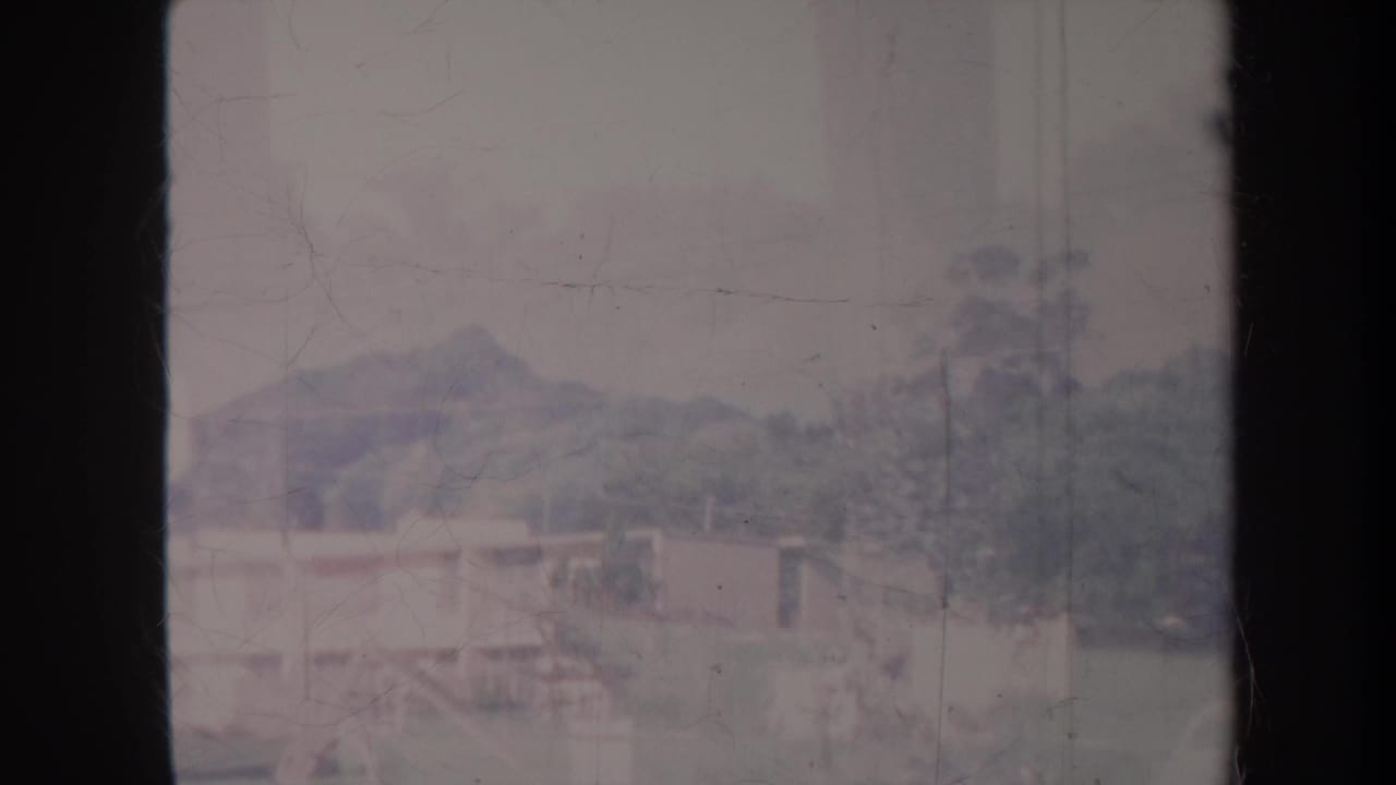 1967 Honolulu home-movie frame showing low buildings, trees, and distant hills under a cloudy sky, with dark 8mm gate edges.