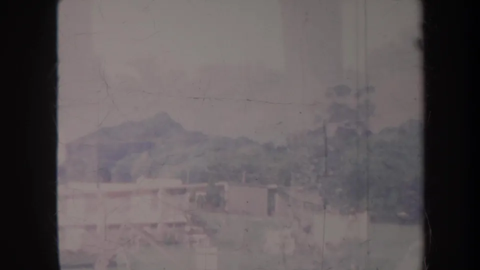 1967 Honolulu home-movie frame showing low buildings, trees, and distant hills under a cloudy sky, with dark 8mm gate edges.