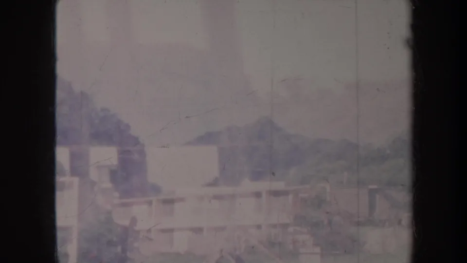 1967 Honolulu frame with pale rooftops, vegetation, and distant hills partly obscured by haze and a vertical 8mm gate shadow.