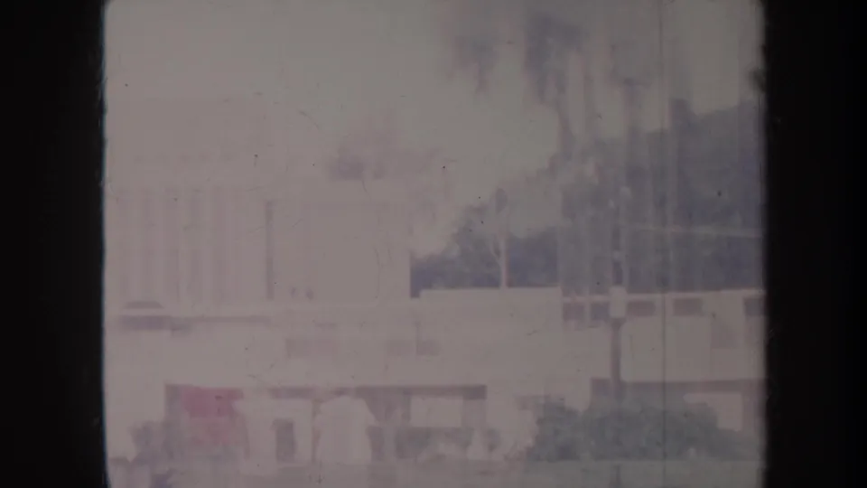1967 Honolulu frame showing pale buildings, dark vegetation, and distant hills through overexposed, hazy 8mm film texture.