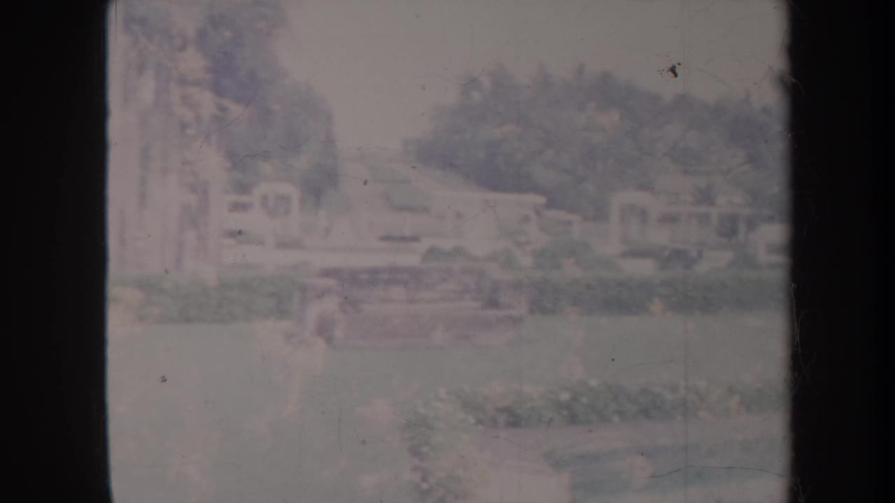 Faded 1967 Honolulu park footage frame with lawn, trees, shrubs, low buildings, and a pale outdoor structure in the background.