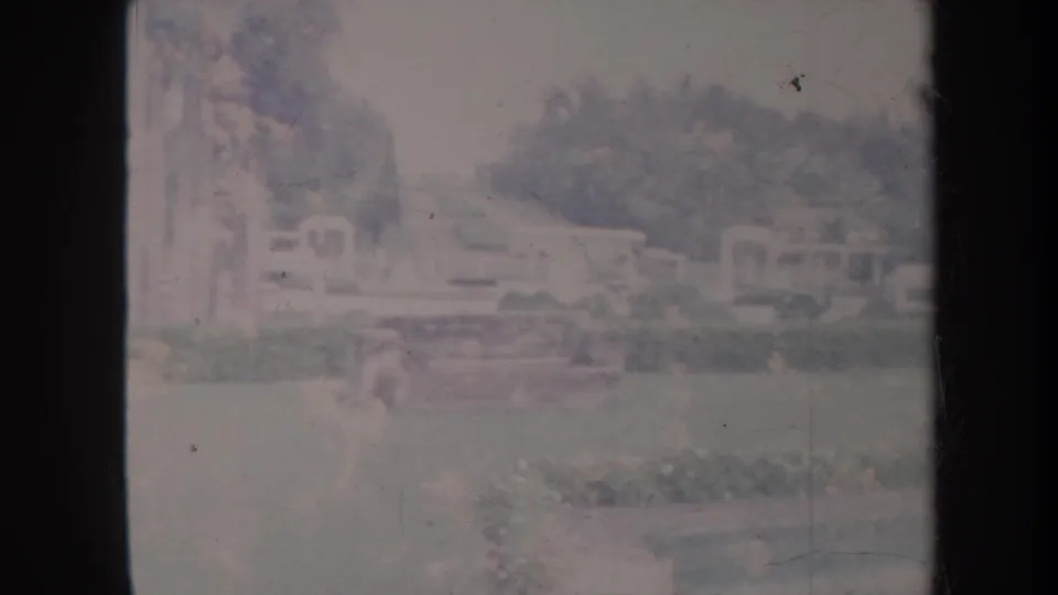 Faded 1967 Honolulu park footage frame with lawn, trees, shrubs, low buildings, and a pale outdoor structure in the background.