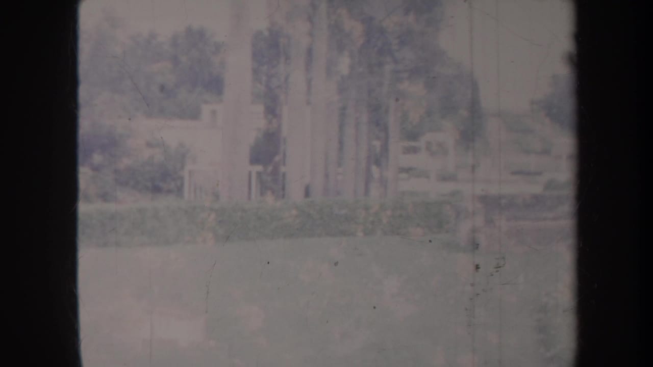 1967 Honolulu home-movie frame with soft-focus pale columns or walls beside lawn, shrubs, and trees in a park-like setting.