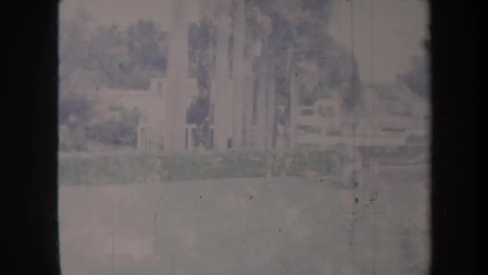 1967 Honolulu home-movie frame with soft-focus pale columns or walls beside lawn, shrubs, and trees in a park-like setting.