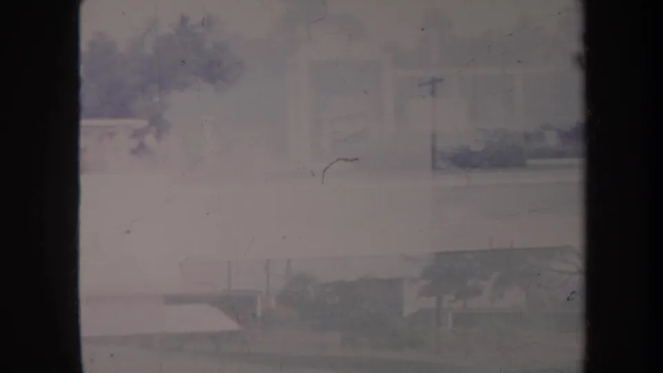 1967 Honolulu frame showing a hazy residential street through a window, with low buildings, utility poles, trees, and parked cars.