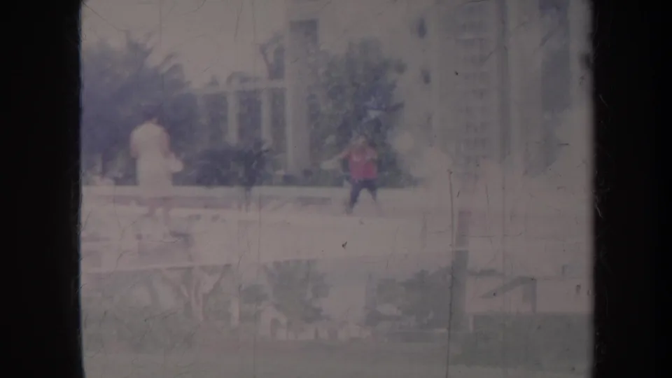 1967 Honolulu suburban frame with two distant pedestrians, houses, trees, a parked car, and dark window mullions in the foreground.