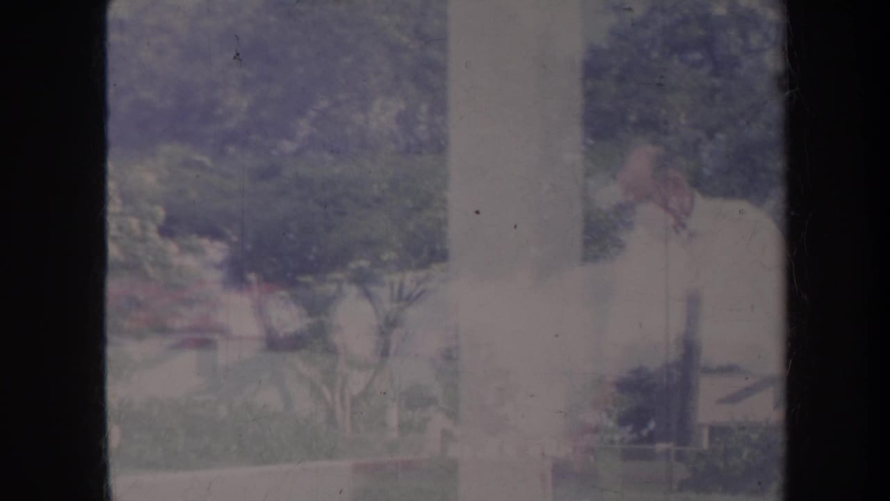 Older man in a light shirt partly obscured by a dark window frame in a garden yard.