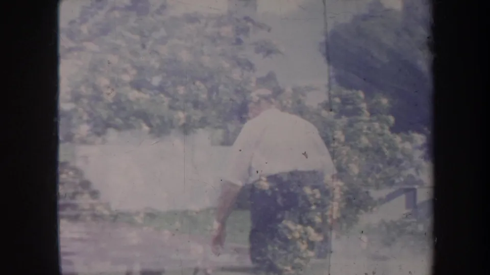 The older man is seen from behind in the 1967 Honolulu yard, moving away through a soft-focus garden view.