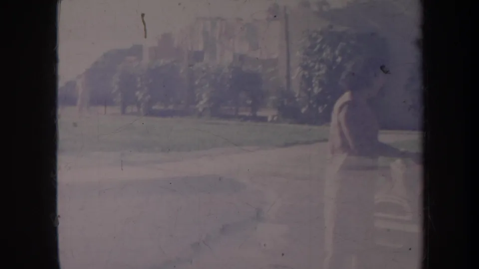 1967 Honolulu home-movie frame showing a person near a parked vehicle beside a lawn and trees, with faded 8mm color.