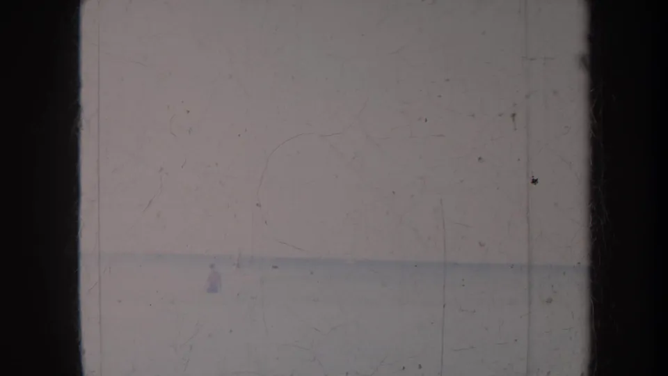 Honolulu home-movie frame looking toward a pale beach or waterline with tiny distant figures and visible film scratches.